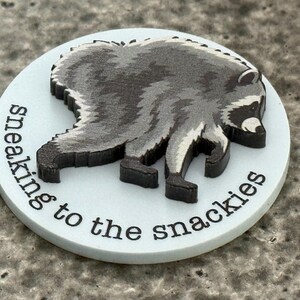 Peut inclure: Un jeton circulaire blanc avec un motif en relief repr&eacute;sentant un raton laveur gris fonc&eacute; et noir en position de marche. Le texte "sneaking to the snackies" est imprim&eacute; en noir, en minuscules et en police sans empattement sur le bord inf&eacute;rieur.