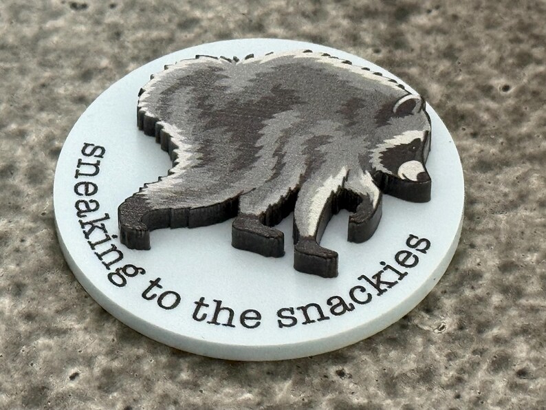 Peut inclure: Un jeton circulaire blanc avec un motif en relief repr&eacute;sentant un raton laveur gris fonc&eacute; et noir en position de marche. Le texte "sneaking to the snackies" est imprim&eacute; en noir, en minuscules et en police sans empattement sur le bord inf&eacute;rieur.