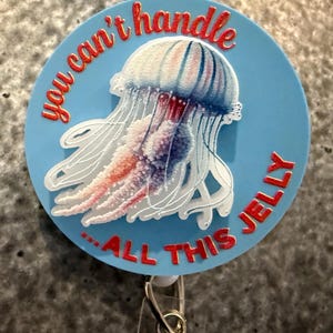 May include: A light blue badge reel with a jellyfish design. The jellyfish is white, red, and blue. The text "you can't handle ALL THIS JELLY" is in red around the edge.