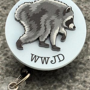 Peut inclure: Un enrouleur de badge circulaire avec un fond bleu clair et une illustration graphique d'un raton laveur gris et noir. Sous le raton laveur, les lettres WWJD sont imprim&eacute;es dans une police sombre, grasse et sans empattement.