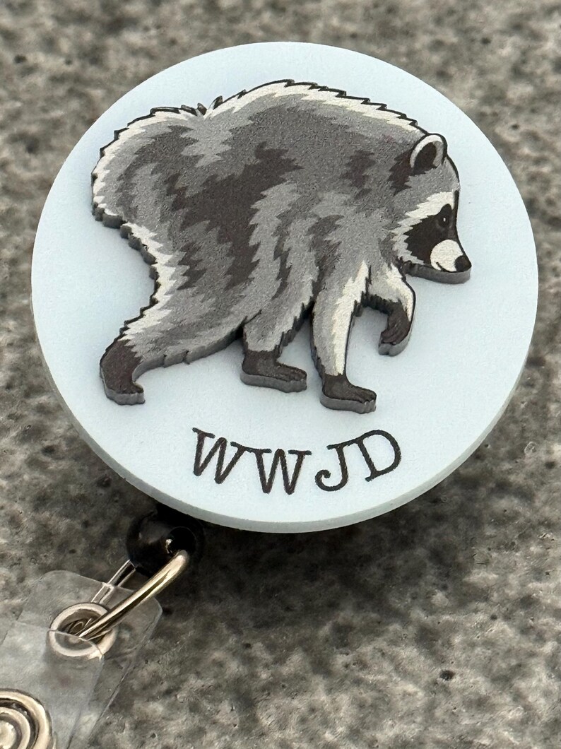 Peut inclure: Un enrouleur de badge circulaire avec un fond bleu clair et une illustration graphique d'un raton laveur gris et noir. Sous le raton laveur, les lettres WWJD sont imprim&eacute;es dans une police sombre, grasse et sans empattement.