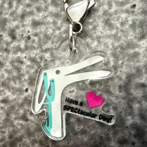 Puede incluir: Un charm de acrílico transparente con un diseño de conejo blanco, detalles turquesa y un corazón rosa. El charm tiene el texto "Have a SPECTacular Day!" y un cierre plateado.