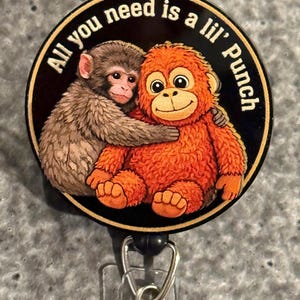 May include: A round badge with a black background and a gold border features two cartoon monkeys hugging. The text "All you need is a lil' Punch" is in a circular pattern around the monkeys. The badge has a retractable reel clip.