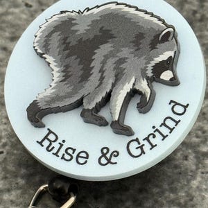 Peut inclure: Un enrouleur de badge circulaire avec un fond bleu clair et un motif de raton laveur gris et noir. Le texte "Rise & Grind" est imprim&eacute; en lettres noires le long de la courbe inf&eacute;rieure du badge. Un clip m&eacute;tallique est fix&eacute; &agrave; la base.