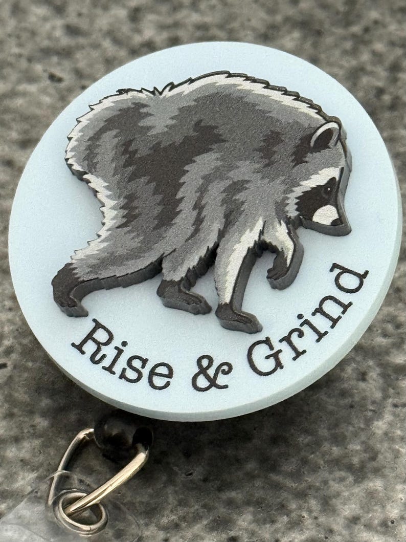 Peut inclure: Un enrouleur de badge circulaire avec un fond bleu clair et un motif de raton laveur gris et noir. Le texte "Rise & Grind" est imprim&eacute; en lettres noires le long de la courbe inf&eacute;rieure du badge. Un clip m&eacute;tallique est fix&eacute; &agrave; la base.