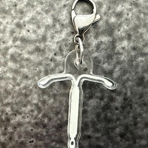 May include: A clear acrylic charm in a T-shape design, attached to a silver-colored lobster clasp. The charm has a white, opaque center and is suspended against a mottled gray background. The charm is a pendant.
