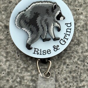 Peut inclure: Un enrouleur de badge circulaire avec un fond bleu clair et une illustration de raton laveur gris et noir. Le texte "Rise & Grind" est imprim&eacute; en noir sur le bord inf&eacute;rieur droit. Une sangle en plastique transparent avec un bouton-pression est fix&eacute;e.