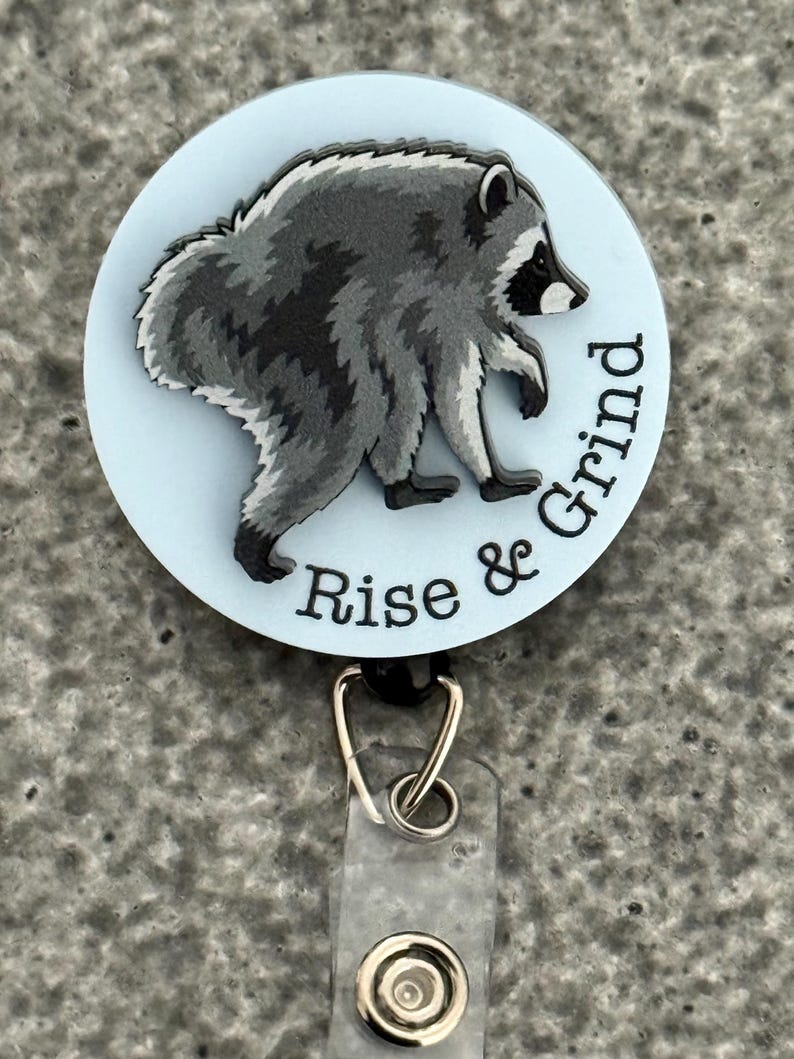 Peut inclure: Un enrouleur de badge circulaire avec un fond bleu clair et une illustration de raton laveur gris et noir. Le texte "Rise & Grind" est imprim&eacute; en noir sur le bord inf&eacute;rieur droit. Une sangle en plastique transparent avec un bouton-pression est fix&eacute;e.