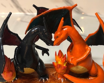 May include: A handcrafted sculpture featuring two Charizard figures, one black and one orange, facing each other. A small flame sculpture is between them. The figures are on a brown base.