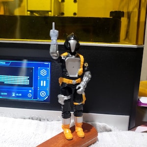 以下が含まれることがあります： ヘルメットと銀色の手をつけた黒と黄色のアクションフィギュアが木製の台座の上に立っています。フィギュアは中指を立てています。