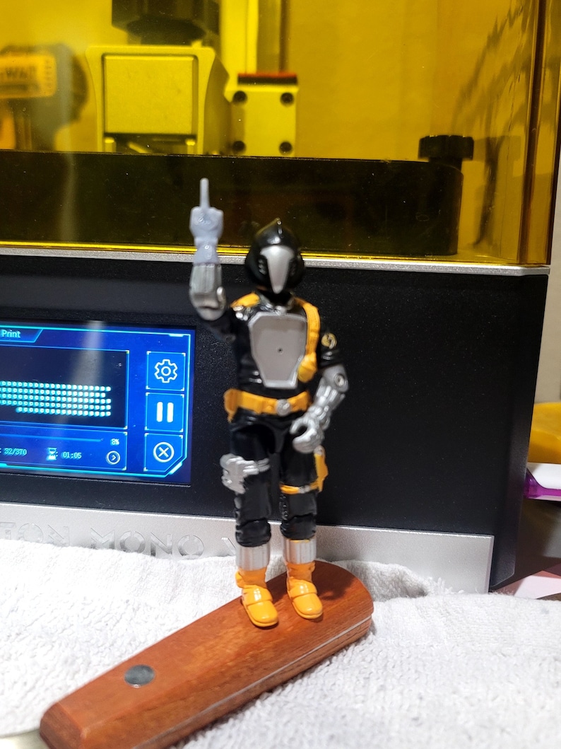 May include: A black and yellow action figure with a helmet and a silver hand, standing on a wooden base. The figure is giving the middle finger.