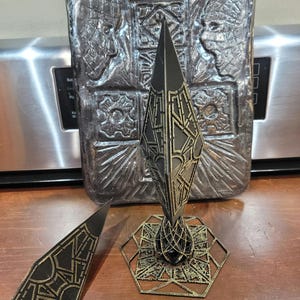 Może przedstawiać: Geometryczny przedmiot dekoracyjny w kolorze czarnym i złotym z ostrym wierzchołkiem i sześciokątną podstawą. Przedmiot jest wykonany z materiału drukowanego w 3D i ma fakturowaną powierzchnię.