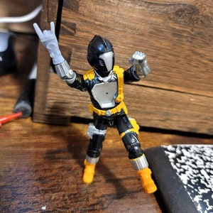 May include: A black and yellow action figure with a silver helmet and a black and silver suit. The figure is posed with one arm raised in a rock and roll hand gesture.
