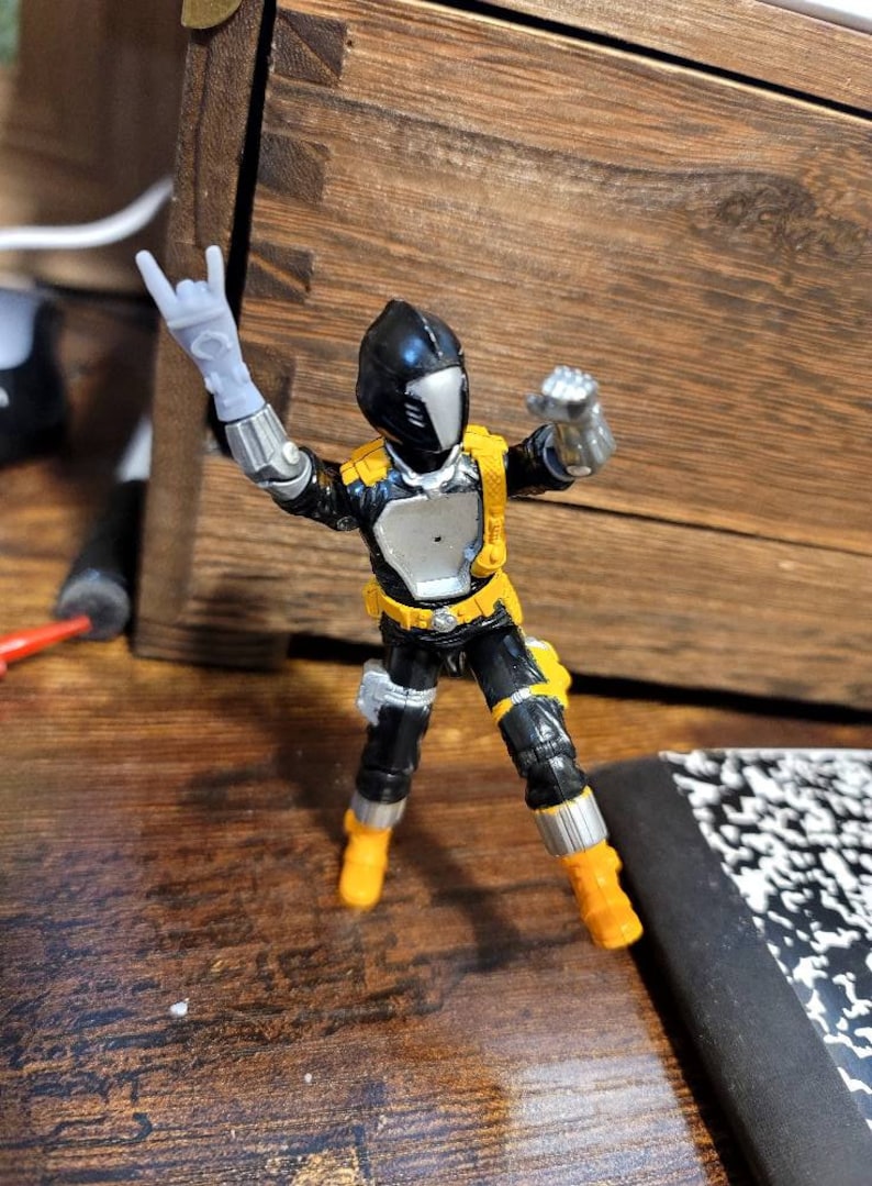 May include: A black and yellow action figure with a silver helmet and a black and silver suit. The figure is posed with one arm raised in a rock and roll hand gesture.