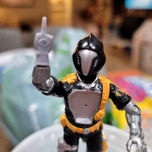 May include: A black and silver action figure with a yellow belt and a black helmet. The figure is holding its right arm up in the air with its middle finger extended.