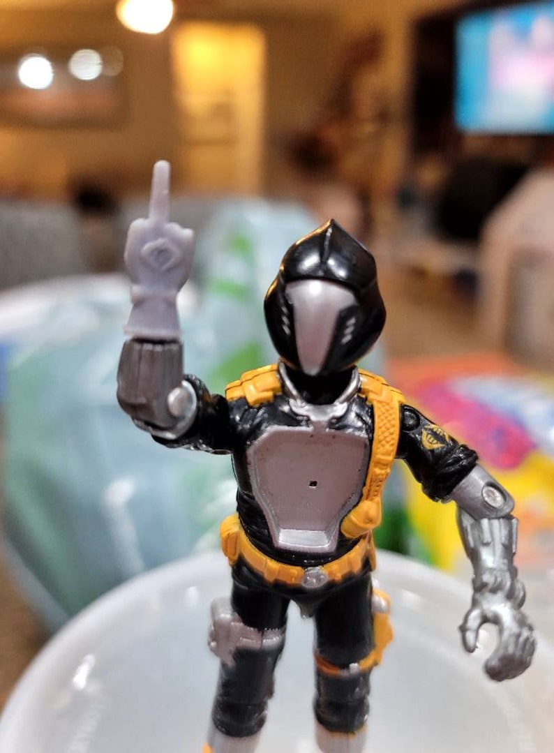May include: A black and silver action figure with a yellow belt and a black helmet. The figure is holding its right arm up in the air with its middle finger extended.