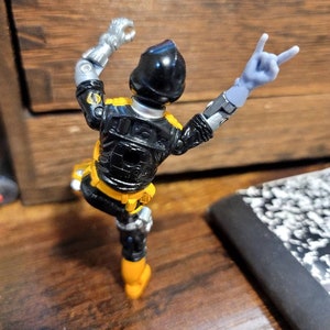 May include: Black and yellow action figure with silver arms and a black helmet. The figure is posed with one arm raised and the other arm making a peace sign.