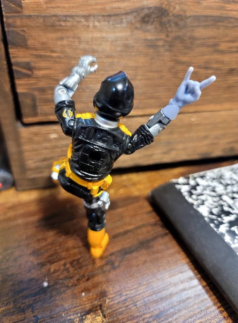 May include: Black and yellow action figure with silver arms and a black helmet. The figure is posed with one arm raised and the other arm making a peace sign.