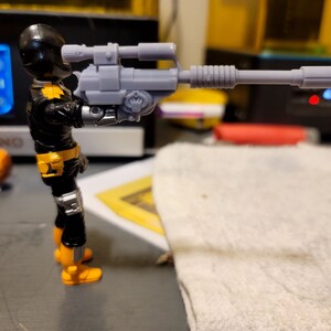 May include: A black and yellow action figure holding a gray plastic rifle with a scope. The figure is wearing a black helmet and a black and yellow suit.
