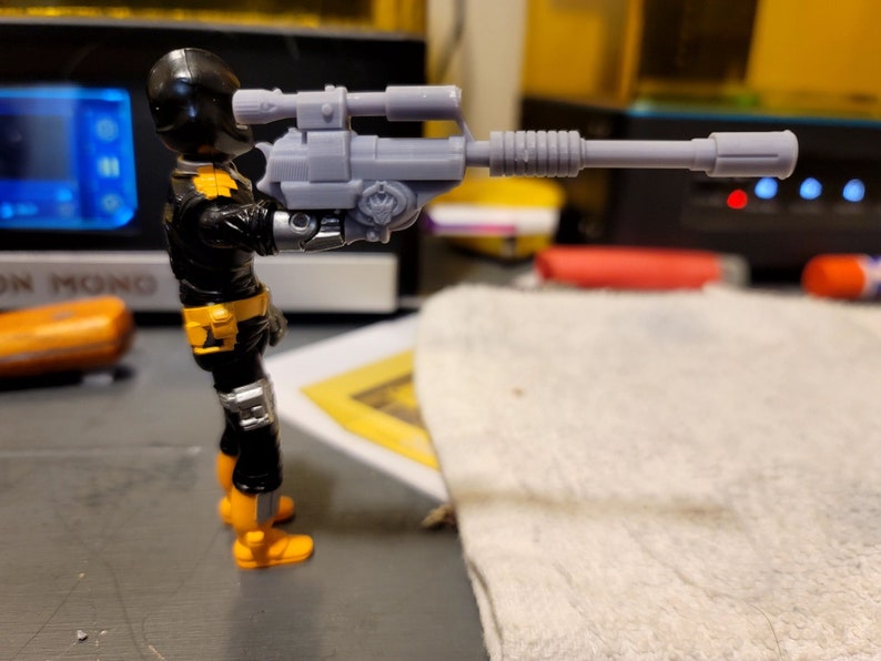 May include: A black and yellow action figure holding a gray plastic rifle with a scope. The figure is wearing a black helmet and a black and yellow suit.