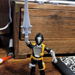 May include: A black and silver action figure with a yellow belt and boots, holding a silver sword. The figure has a black helmet with a silver visor.