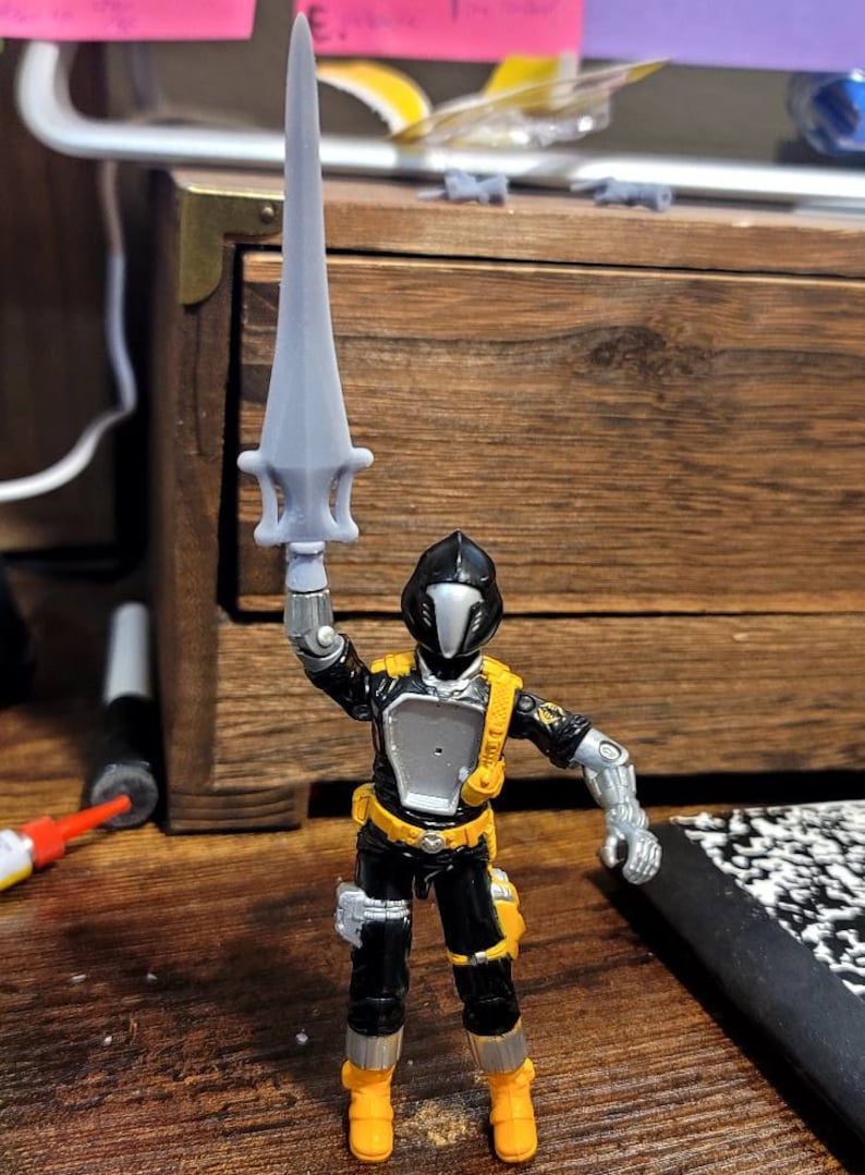 May include: A black and silver action figure with a yellow belt and boots, holding a silver sword. The figure has a black helmet with a silver visor.