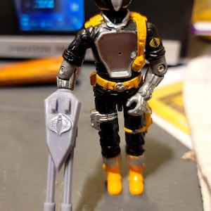 May include: A black and silver action figure with a yellow belt and boots. The figure is holding a gray weapon with a Cobra logo on it.