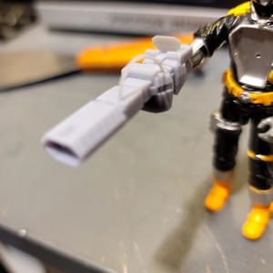 May include: A gray plastic toy gun with a black handle and a silver barrel. The gun is held by a toy action figure.