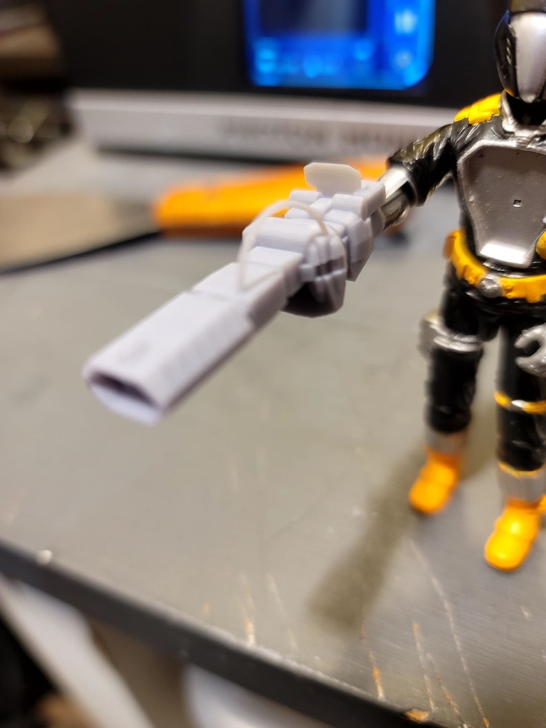 May include: A gray plastic toy gun with a black handle and a silver barrel. The gun is held by a toy action figure.