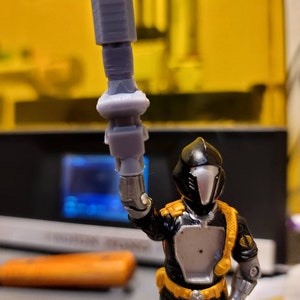 May include: A black and silver action figure with a black helmet and a gray weapon raised in the air. The figure is wearing a yellow belt and has a black and yellow Cobra logo on the shoulder.
