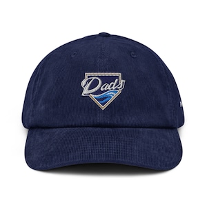 Peut inclure: Une casquette de baseball en velours côtelé bleu marine avec un logo brodé qui dit "Dad's" en écriture cursive blanche avec un motif de vague bleue à l'intérieur d'un losange de baseball beige.