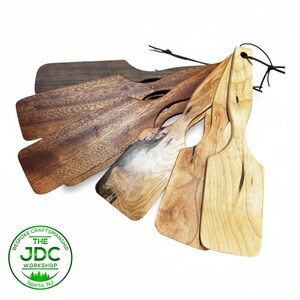 May include: A collection of handcrafted wooden spatulas in various shades of brown, from dark to light. Each spatula features a unique grain pattern and a handle with a hole for hanging. The JDC Workshop logo is visible in the lower left corner.