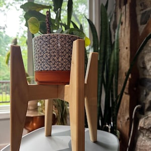 May include: A wooden plant stand with a light finish supports a patterned ceramic pot. The pot has a geometric design in black and white, with an orange base. The stand is on a gray surface, with plants visible in the background.
