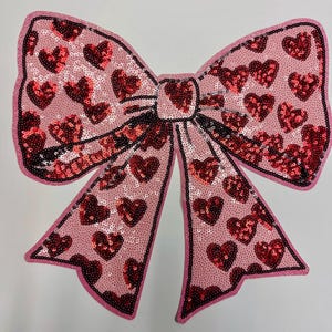 May include: A pink fabric bow with red sequins in the shape of hearts. The bow is a decorative embellishment for clothing or crafts.