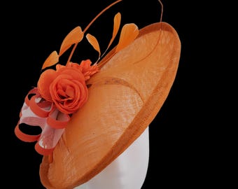 Op de afbeelding: Oranje fascinator hoed met een grote roos en decoratieve veren. De hoed heeft een brede rand en is gemaakt van geweven materiaal. Een wit en oranje lint is aan de zijkant bevestigd. Gepresenteerd op een witte mannequin hoofd.