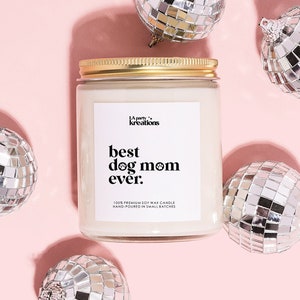 May include: A white candle with a gold lid and a label that reads "best dog mom ever." The candle is surrounded by silver disco ball ornaments on a pink background.