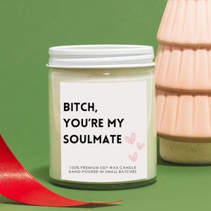 Pode incluir: Uma vela de cera de soja branca com um r&oacute;tulo preto que diz "BITCH, YOU'RE MY SOULMATE" com cora&ccedil;&otilde;es rosa. A vela est&aacute; sobre uma superf&iacute;cie verde com uma fita vermelha e uma &aacute;rvore de Natal de cer&acirc;mica rosa.