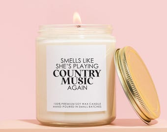 May include: A lit, white soy wax candle in a glass jar with a gold lid. The label reads "Smells Like She's Playing Country Music Again." The candle sits on a peach-colored block against a pink background.