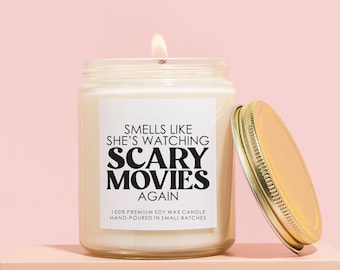 May include: A white soy wax candle with a gold lid. The candle label reads "Smells like she's watching scary movies again." 100% premium soy wax candle, hand-poured in small batches.