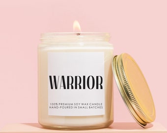 May include: A white soy wax candle in a clear glass jar with a gold lid. The candle is lit and the label reads "WARRIOR 100% PREMIUM SOY WAX CANDLE HAND-POURED IN SMALL BATCHES". The candle is sitting on a light pink surface.