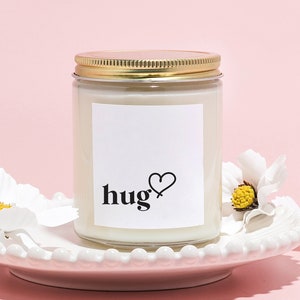 May include: A white candle in a glass jar with a gold lid. The jar has a white label with the word "hug" and a heart drawn on it. The candle is sitting on a white plate with white flowers around it.