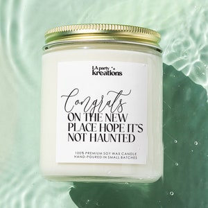 May include: A white soy wax candle with a gold lid. The label reads "A party kreations" and "Congrats on the new place hope it's not haunted". The candle is floating in a pool of water.