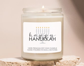 May include: A white soy wax candle with a label that says "happy hanukkah" and features a silver menorah with gold flames. The candle is hand-poured in small batches.