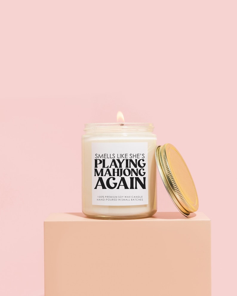 K&ouml;nnte beinhalten: Eine wei&szlig;e Sojawachskerze mit einem schwarzen Etikett, auf dem "Smells like she's playing mahjong again" steht. Die Kerze befindet sich in einem durchsichtigen Glasgef&auml;&szlig; mit einem goldenen Deckel. Die Kerze steht auf einer hellrosa Oberfl&auml;che.