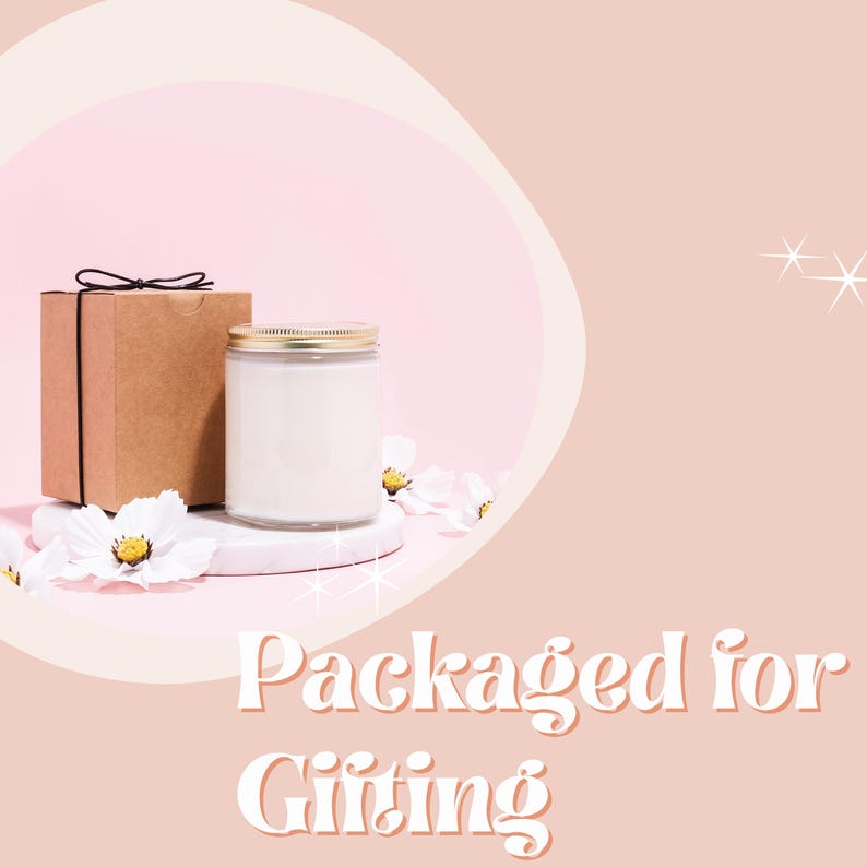May include: A white candle in a glass jar with a gold lid, packaged in a brown box with a black ribbon. The candle is on a white pedestal with white flowers. The background is a light pink with a white and beige abstract shape. The text "Packaged for Gifting" is at the bottom of the image.