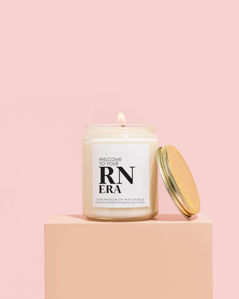 May include: A white soy wax candle with a gold lid. The candle is labeled "Welcome to your RN Era" and "100% Premium Soy Wax Candle Hand-Poured in Small Batches".