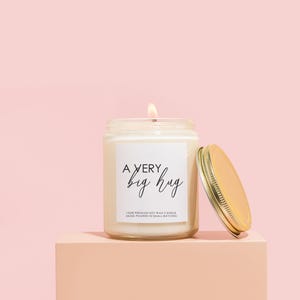 Pode incluir: Uma vela de cera de soja branca em um pote de vidro transparente com tampa dourada. A vela tem um r&oacute;tulo que diz "A VERY big hug" e "100% PREMIUM SOY WAX CANDLE HAND-POURED IN SMALL BATCHES".
