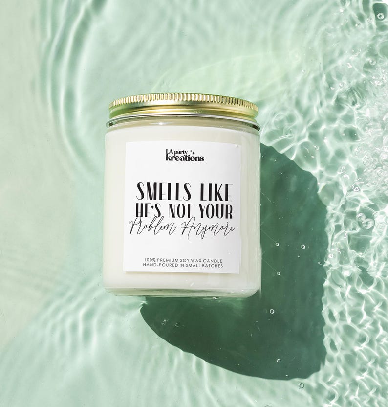 May include: White soy wax candle with a gold lid. The label reads "LA party kreations" and "SMELLS LIKE HE'S NOT YOUR Problem Anymore". The candle is floating in a pool of water.