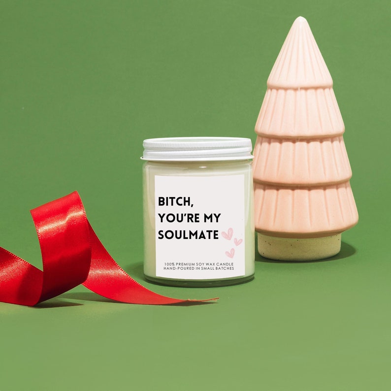 Pode incluir: Uma vela de cera de soja branca com um r&oacute;tulo preto que diz "BITCH, YOU'RE MY SOULMATE" com cora&ccedil;&otilde;es rosa. A vela est&aacute; sobre uma superf&iacute;cie verde com uma fita vermelha e uma &aacute;rvore de Natal de cer&acirc;mica rosa.