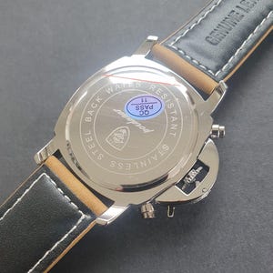 May include: The back of a silver watch with a black leather band. The watch face says "Water Resistant" and "Stainless Steel Back". There is a blue sticker on the watch that says "QC Pass 11".